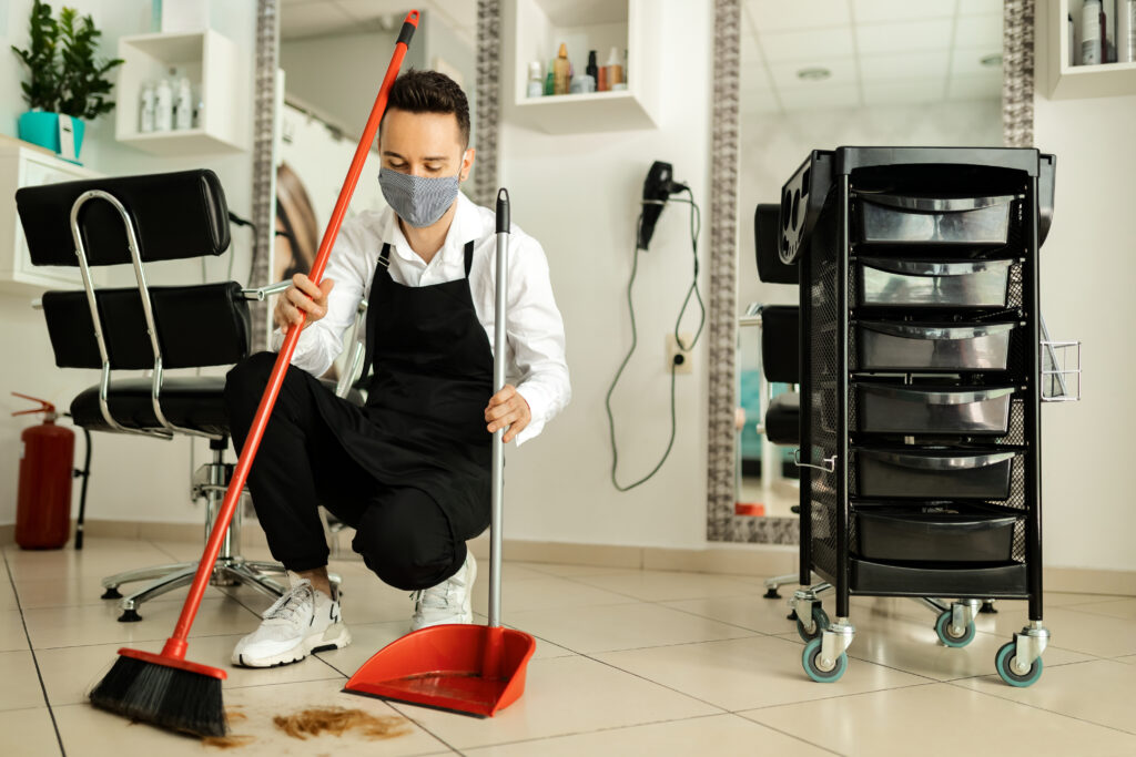 hairdresser with protective face mask cleaning salon after haircut